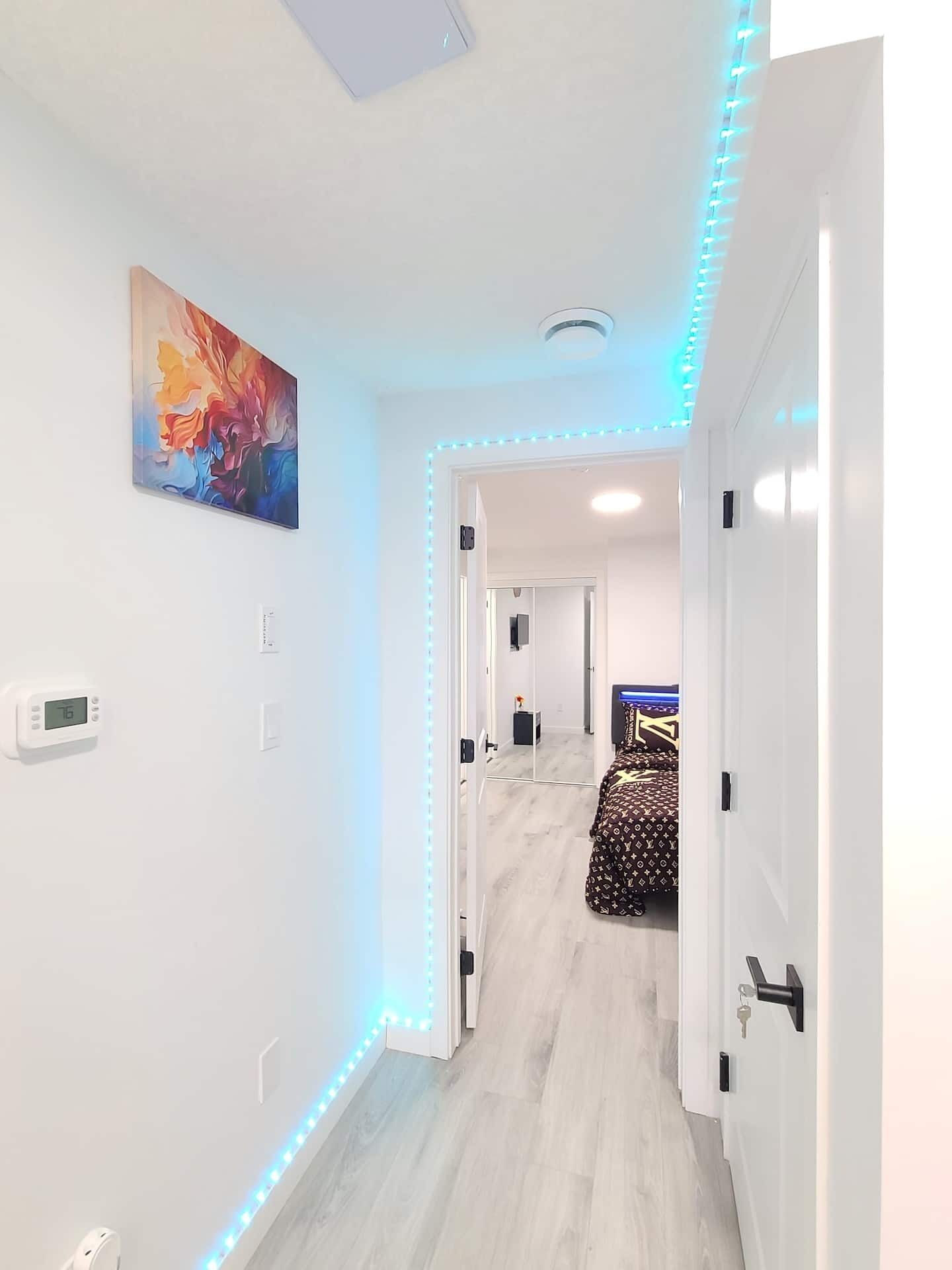 Um corredor com paredes brancas, uma luminária no teto, um quadro e uma cama com colcha estampada.