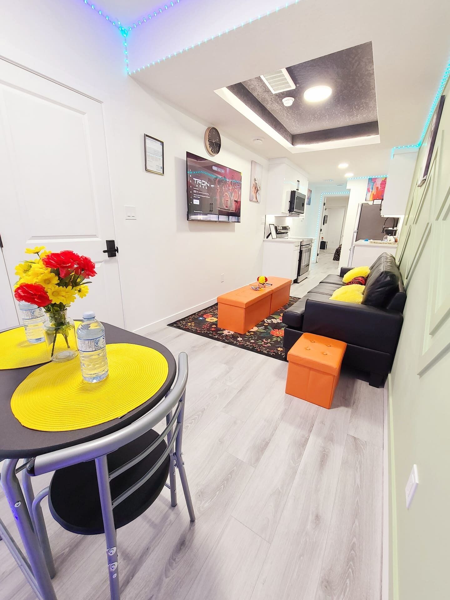 A modern living room with a dining area, featuring a round table with yellow placemats, a sofa with orange cushions, and a TV mounted on the wall.