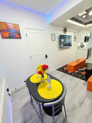 A modern living room with a dining area, a flat-screen TV mounted on the wall, and a kitchen in the background. - SwitHome Properties Cher Cozy Suite (Pond View), Near YEG Airport. (Edmonton)