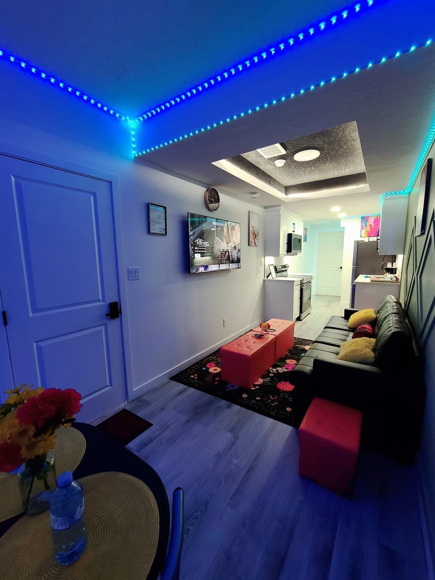 Ein modernes Wohnzimmer mit einer Couch, einem Couchtisch und einem Fernseher. Im Hintergrund befindet sich eine Küchenzeile mit einem Kühlschrank und einer Mikrowelle.