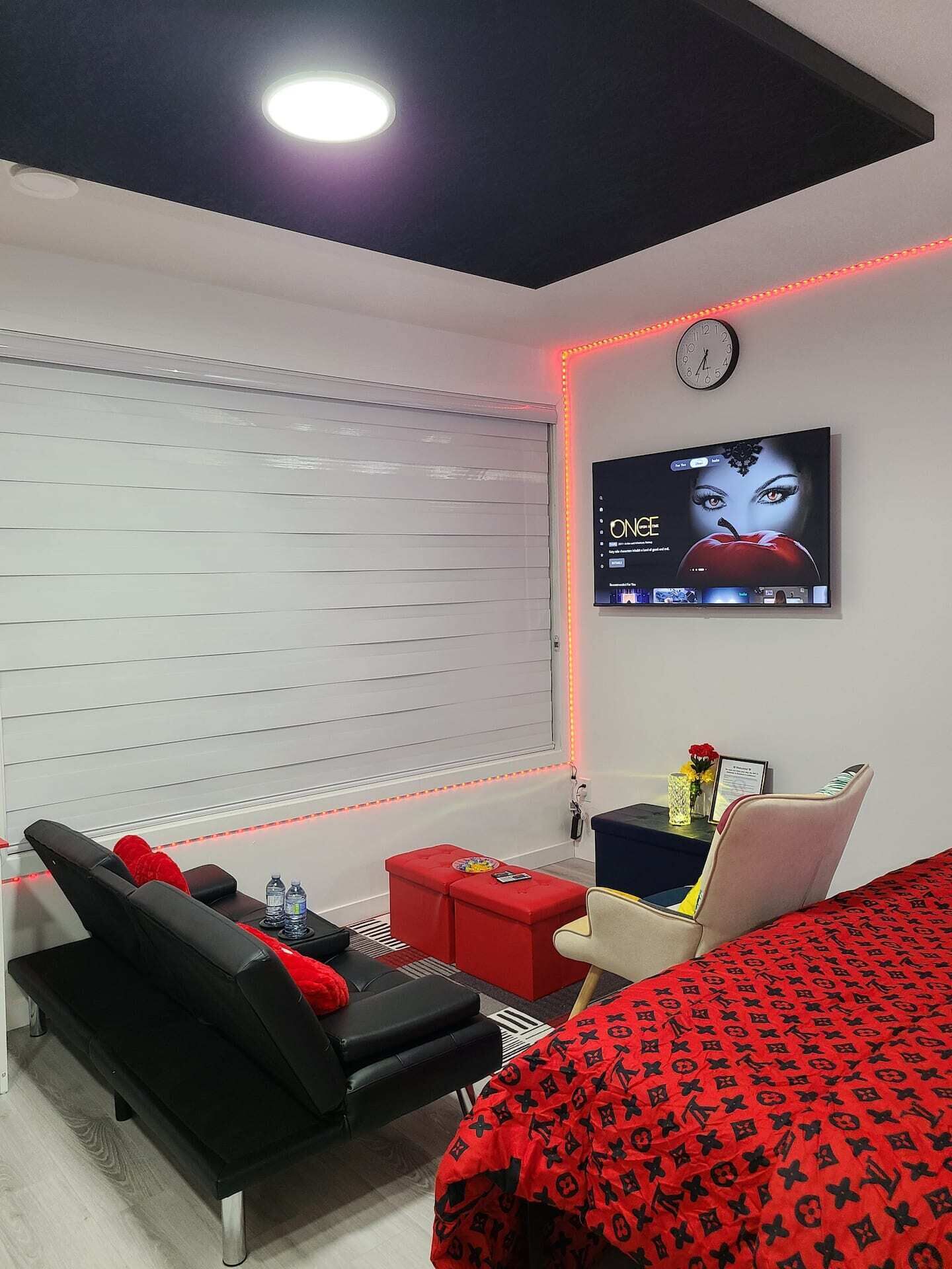 A modern hotel room with a flat-screen TV mounted on the wall, a sofa, a red ottoman, and a bed with a red patterned bedspread.