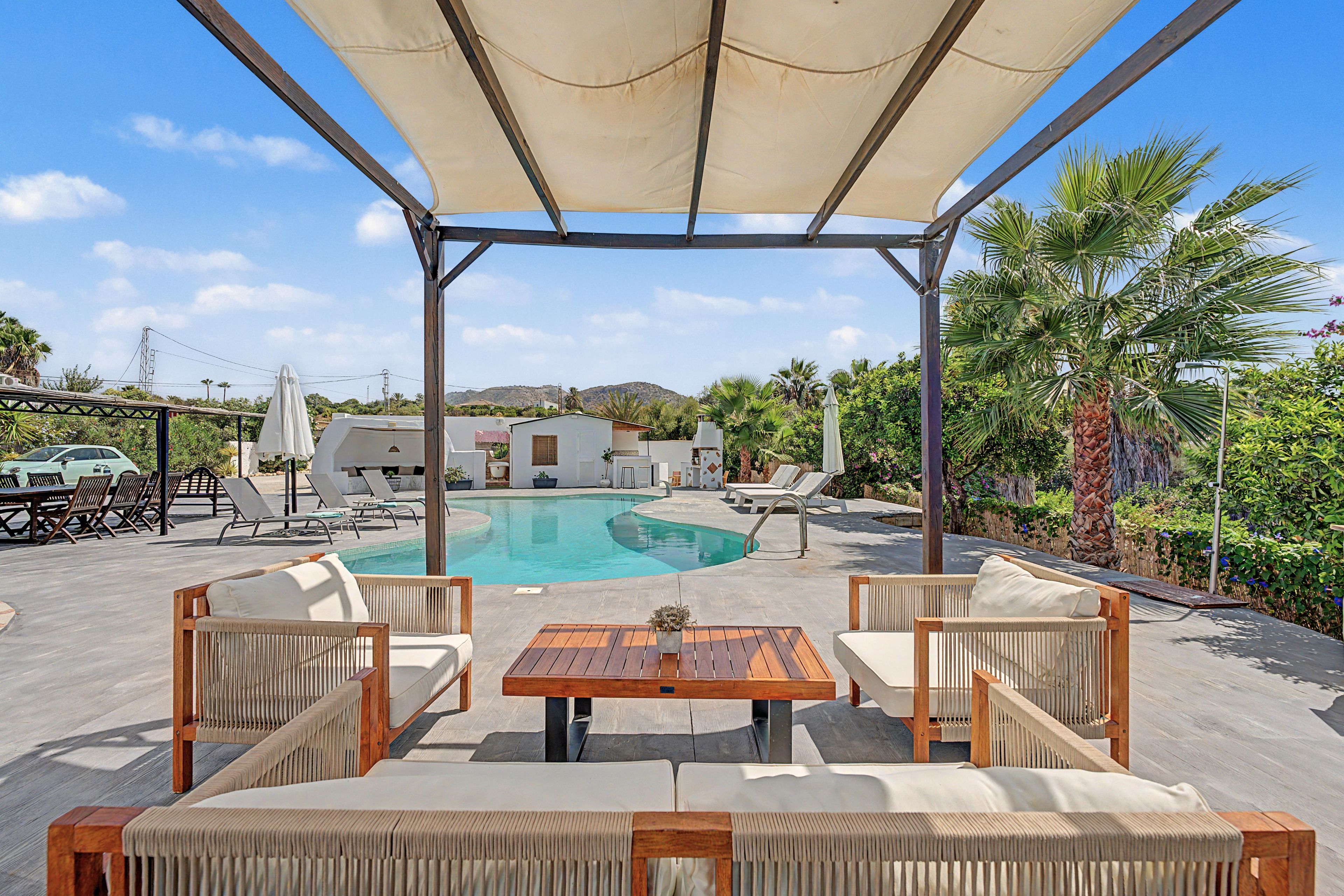 Zona de piscina con sillones de descanso y una mesa bajo una pérgola.