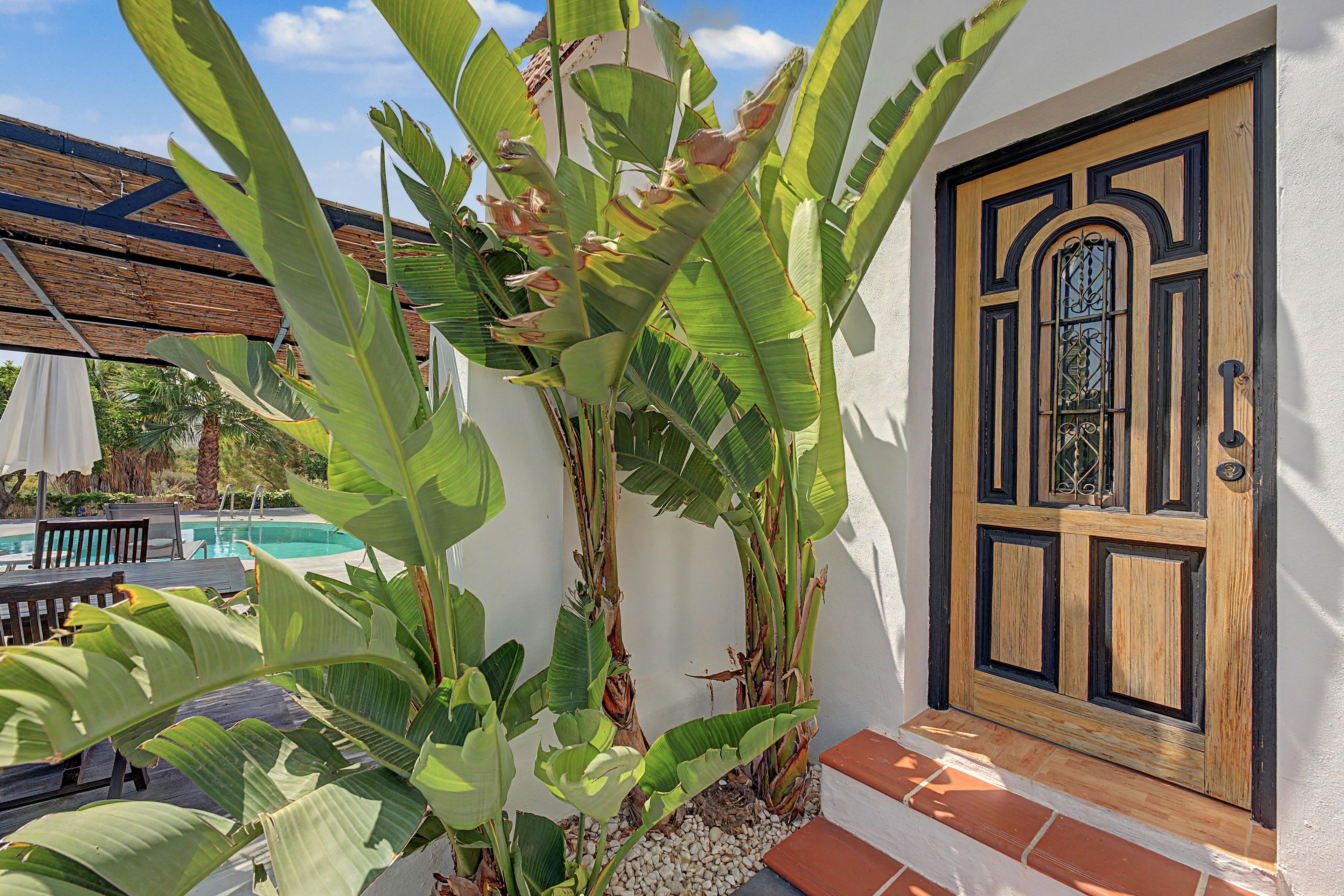 Una casa con puerta de madera, piscina y grandes plantas de banano.