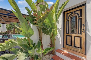Una casa con puerta de madera, piscina y grandes plantas de banano.