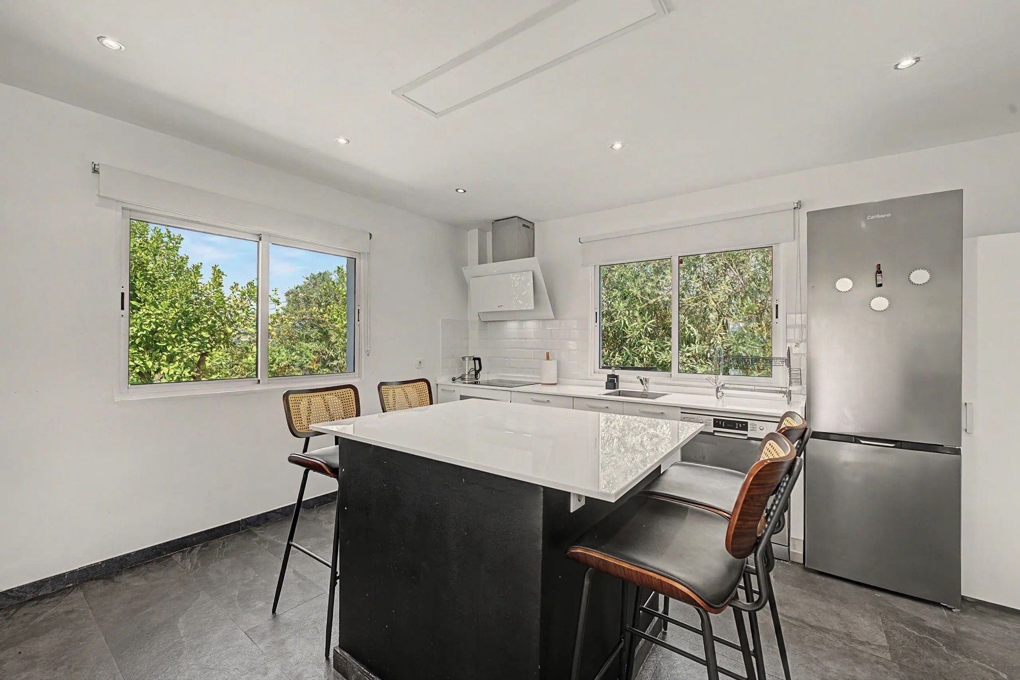 Una cocina moderna con una isla central, taburetes de bar, un refrigerador y ventanas amplias.