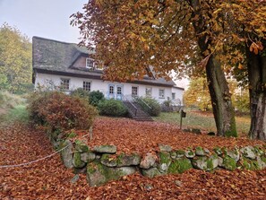 Un edificio blanco con tejado de paja, rodeado de un muro de piedra y un árbol con hojas otoñales.
