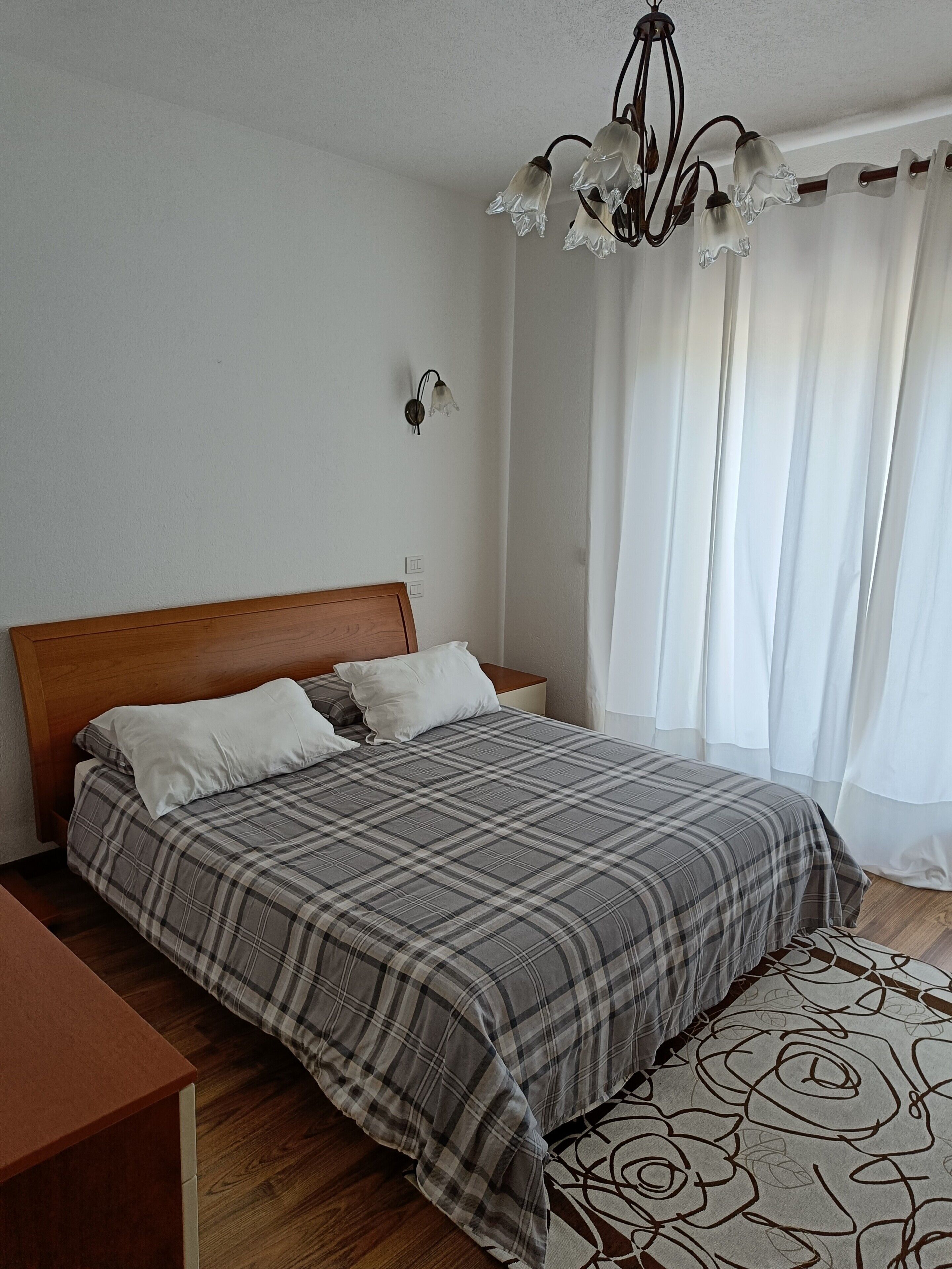 A bedroom with a wooden headboard, a bed with a plaid bedspread, a bedside table, a wall-mounted light fixture, and a window with sheer curtains.