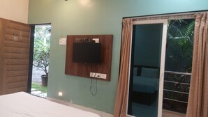 A room with a bed, a television mounted on a wooden panel, a sliding glass door with curtains, and a small window with a view of greenery. - Seashell Retreat (Shrivardhan)