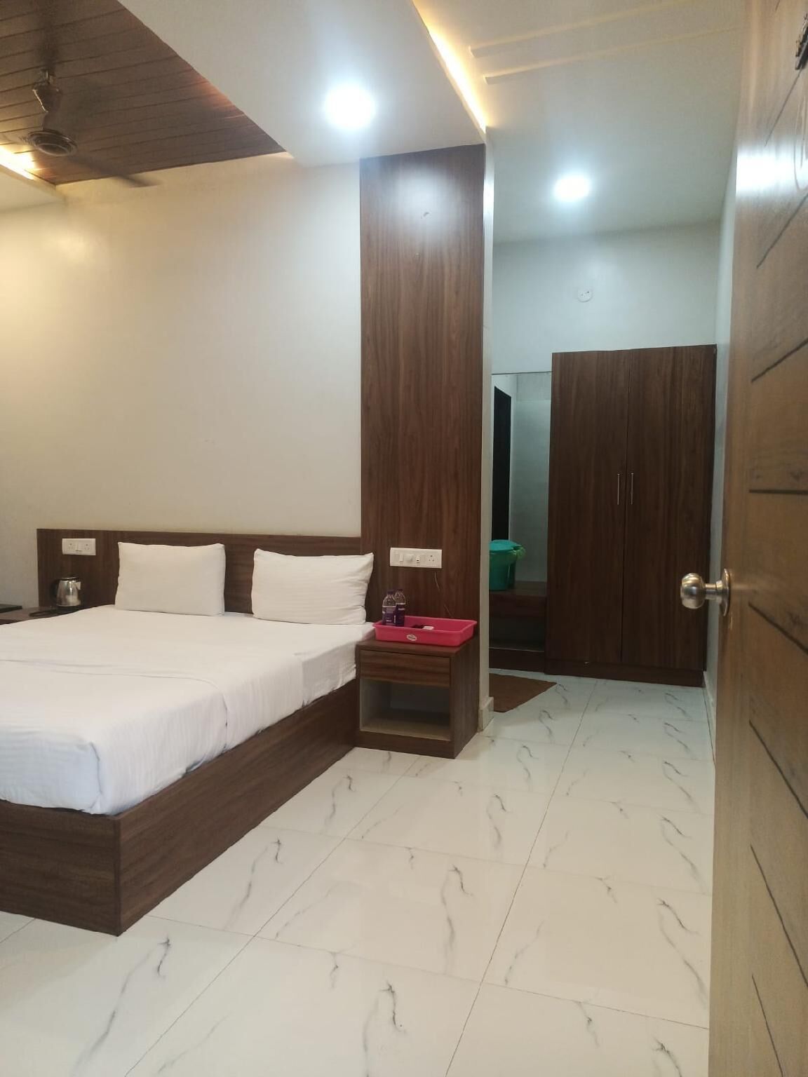 Een hotelkamer met een bed, nachtkastje, houten garderobe en een zichtbare badkamer door een open deur.