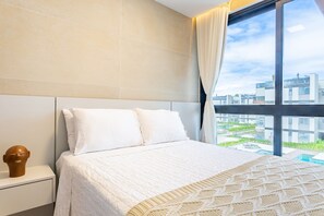 A hotel room with a large bed, a nightstand, a window with curtains, and a view of buildings and greenery outside. - Experiência Exclusiva em Muro Alto  (N. Sra. do Ó)