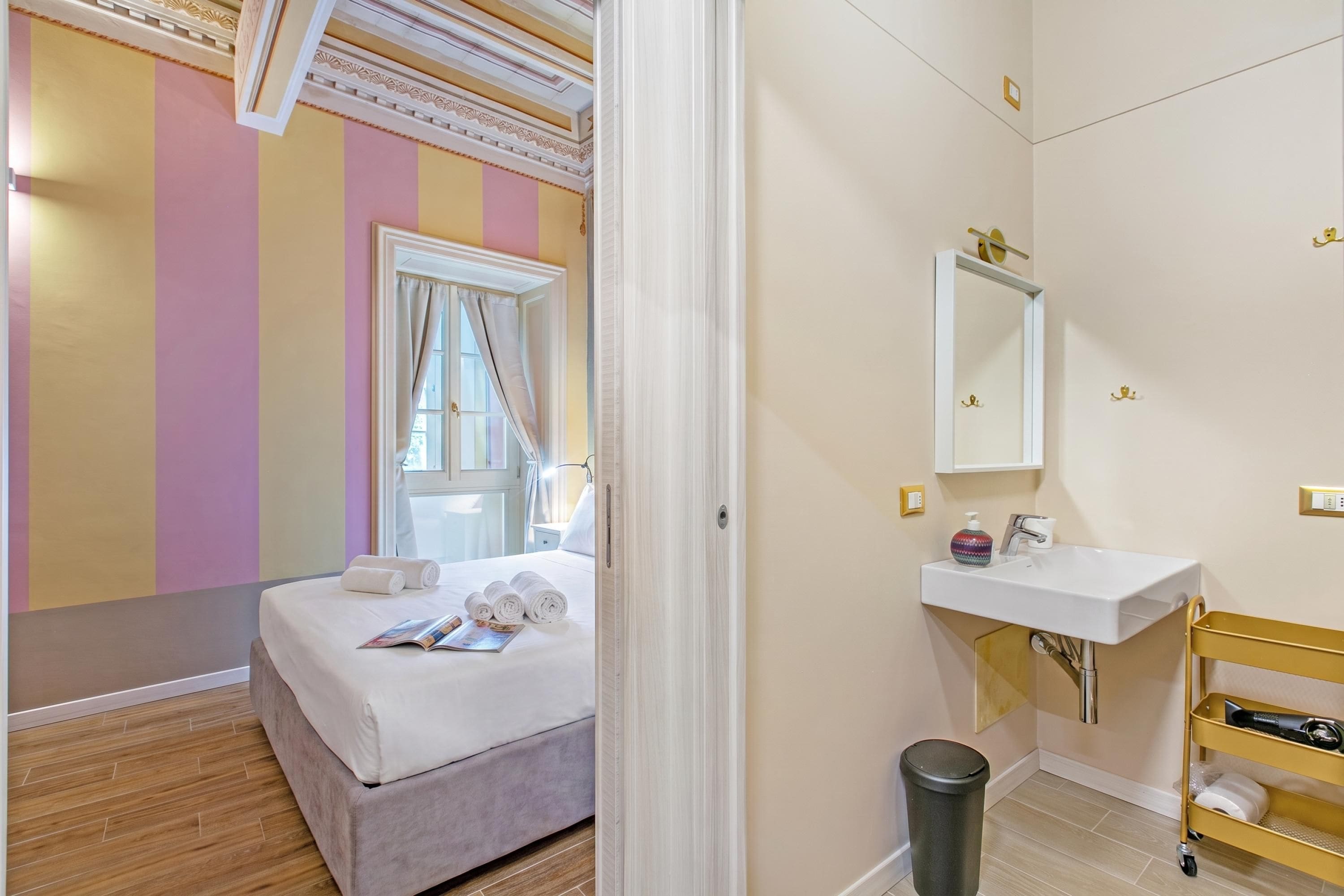 Ett hotellrum med en säng, ett fönster med gardiner, ett sängbord, en spegel, ett handfat och en papperskorg.