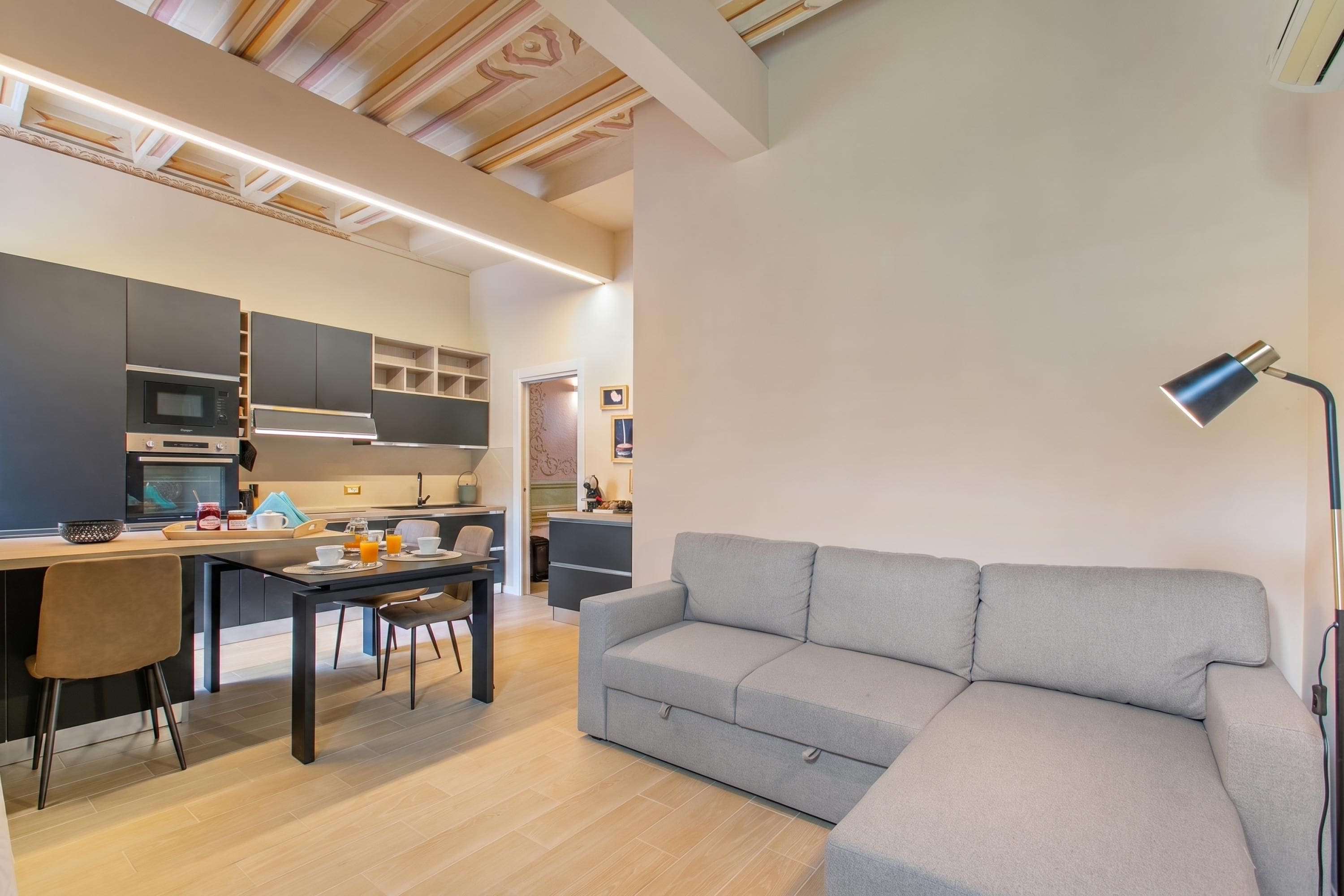 Una cucina moderna con elettrodomestici integrati, un'area pranzo con tavolo e sedie, e un divano grigio in un soggiorno.