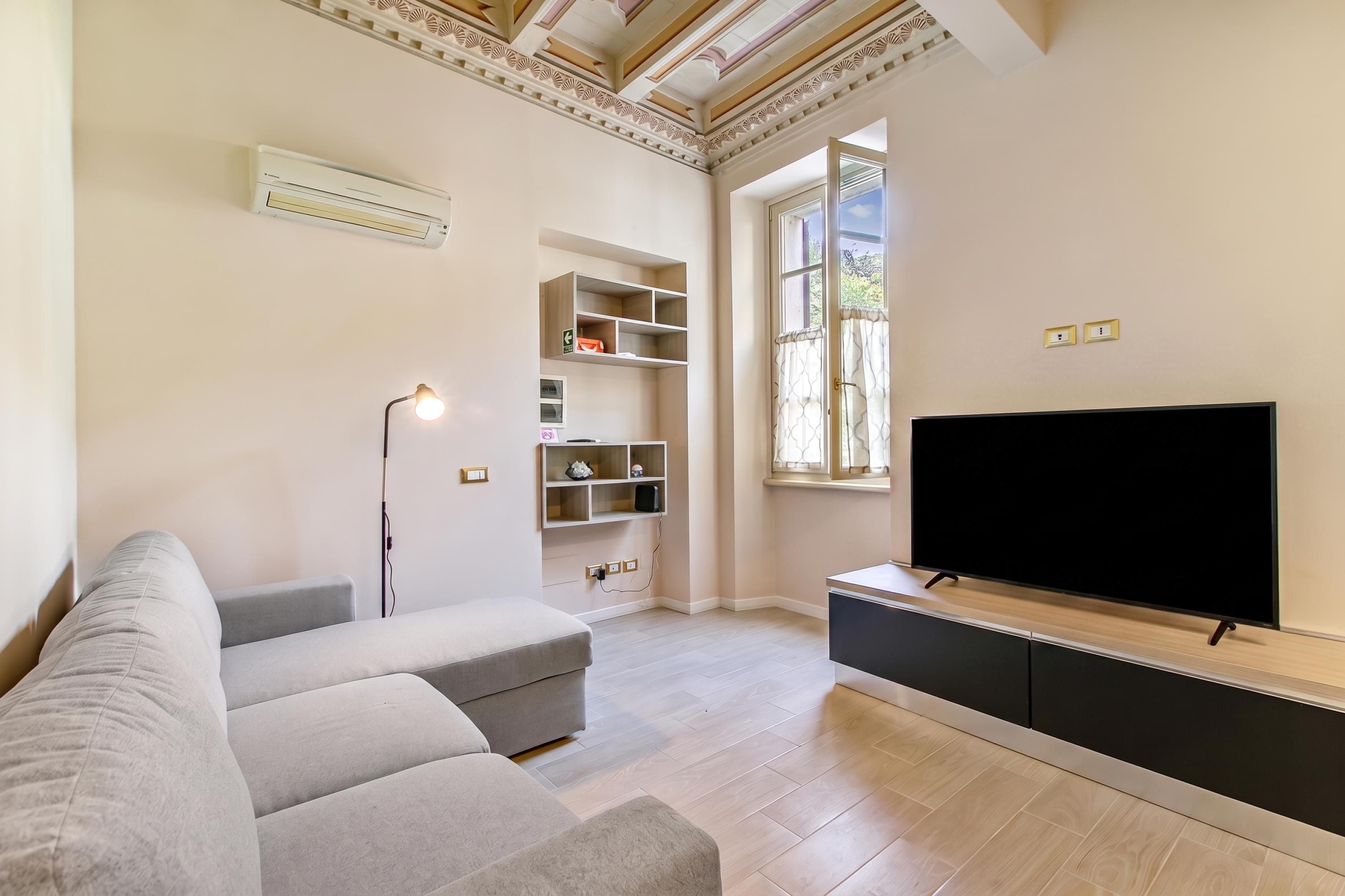 Una sala de estar con un sofá gris, un televisor de pantalla plana sobre un soporte de madera, una lámpara de pie y estanterías empotradas.