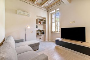 Een woonkamer met een grijze bank, een flatscreen televisie op een houten standaard, een vloerlamp en inbouwplanken.