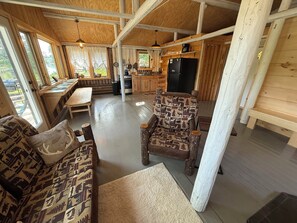 A rustic cabin interior with a wooden dining table, a patterned sofa, a unique chair, and a kitchen area with a refrigerator.