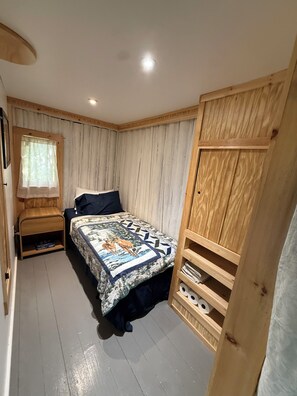 A bedroom with a bed, a wooden headboard, a window with curtains, and a shelf with books and toiletries.