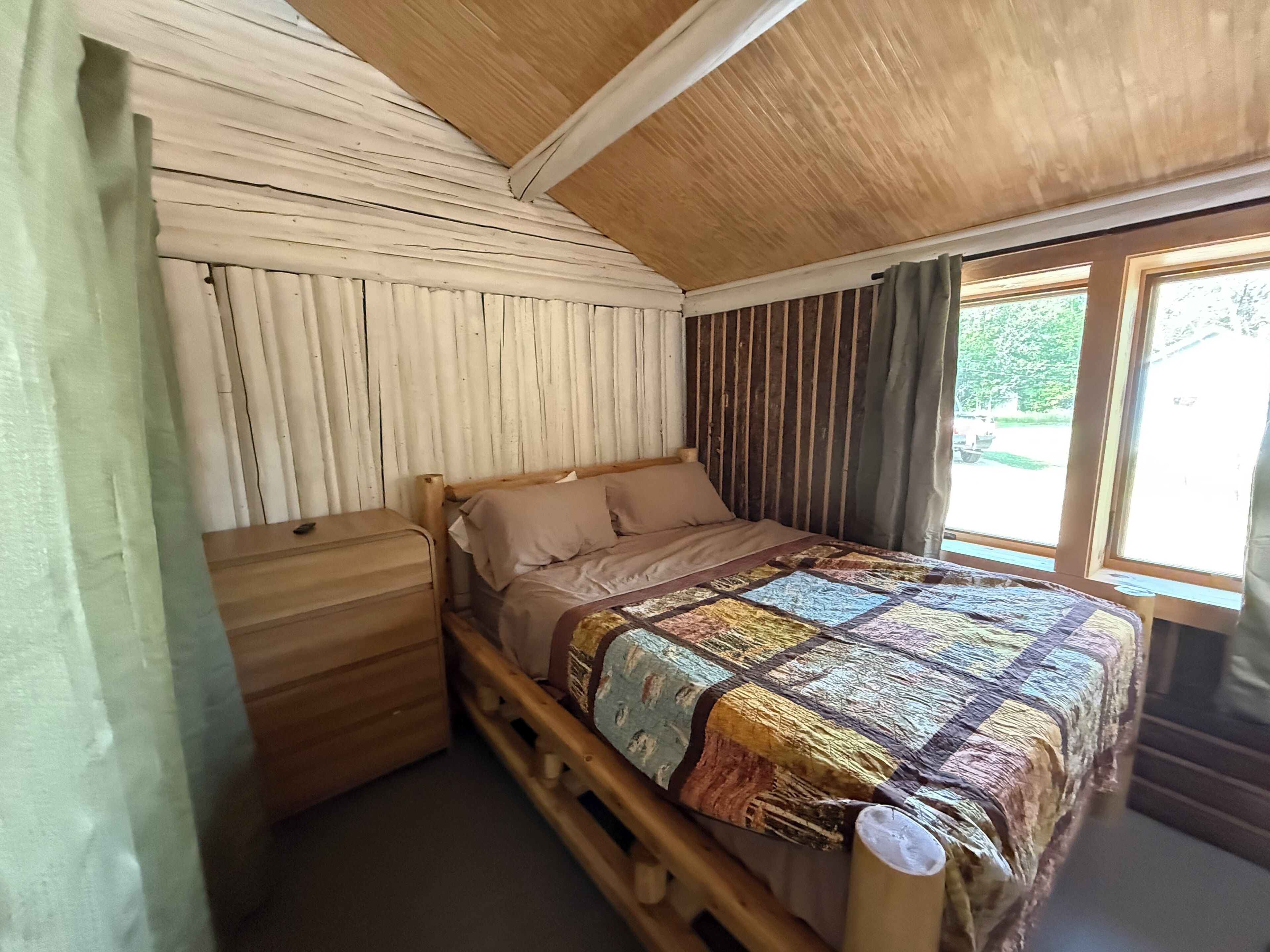 Ein hölzernes Bett mit Patchworkdecke, ein Nachttisch aus Holz und ein Fenster mit Vorhängen.
