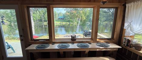 Puinen sisustus, jossa on pöytä ja kolme ikkunaa, joista avautuu näkymä järvelle.