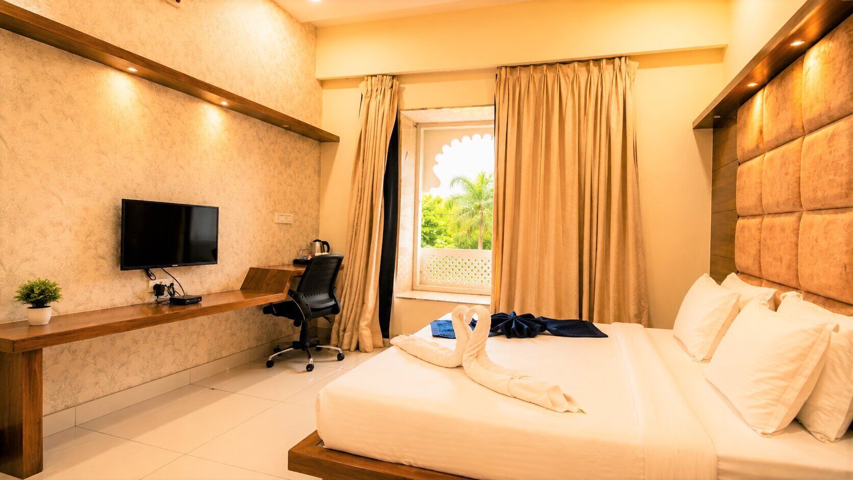 Habitación de hotel con cama, escritorio, silla, televisión y ventana con cortinas.