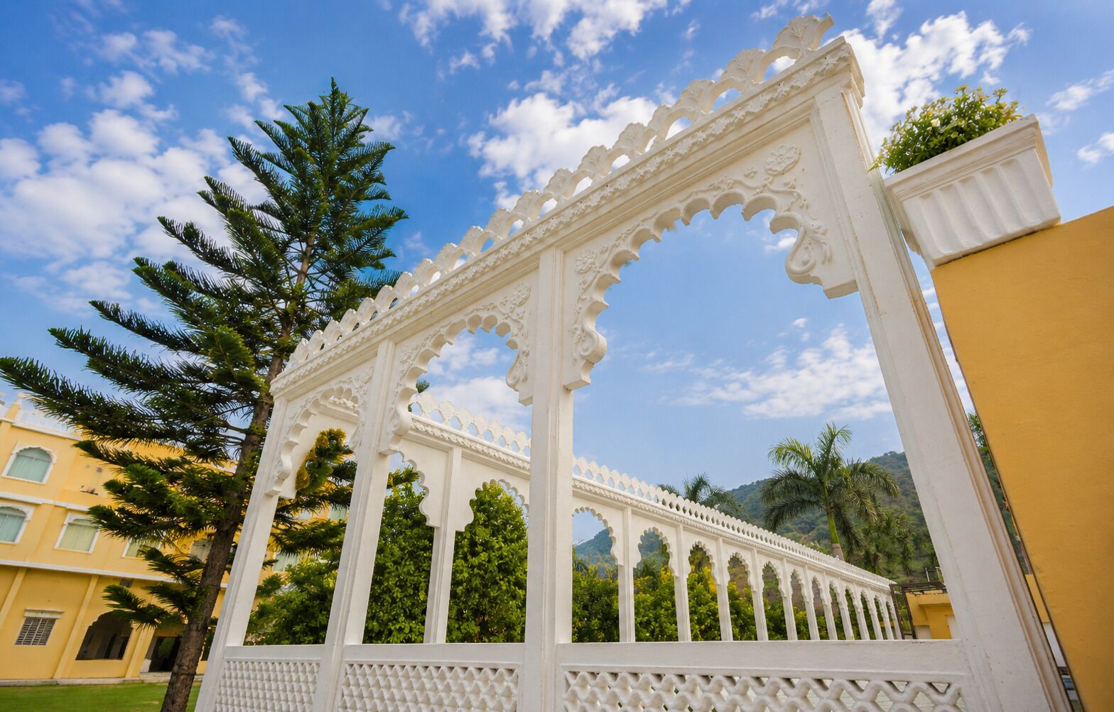 Una estructura blanca ornamentada con intrincados grabados, rodeada de vegetación y un cielo despejado.