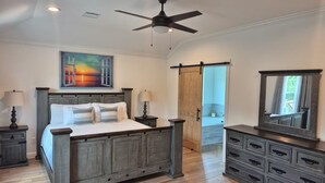 Een slaapkamer met een bed, nachtkastjes, een dressoir, een plafondventilator, een schilderij en een schuifdeur die naar een andere kamer leidt.