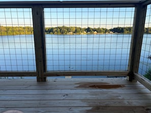 A wooden deck with a view of a calm lake and a forested shoreline. - Custom Lake House get-a-way.  Tranquil 4-bedroom house with AC on Crispell Lake (Clarklake)