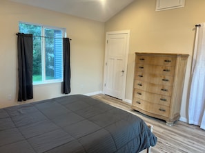 A bedroom with a bed, a wooden dresser, a window with curtains, and a door.