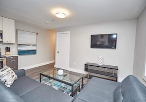 A modern living room with a glass coffee table, grey sofas, a flat-screen TV mounted on the wall, and a kitchen area with cabinets and a painting. - This condo is perfect for families, friend groups and couples. (NIAGARA FALLS)