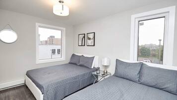 A bedroom with two beds, a bedside table with a lamp, and a window with a view of buildings.