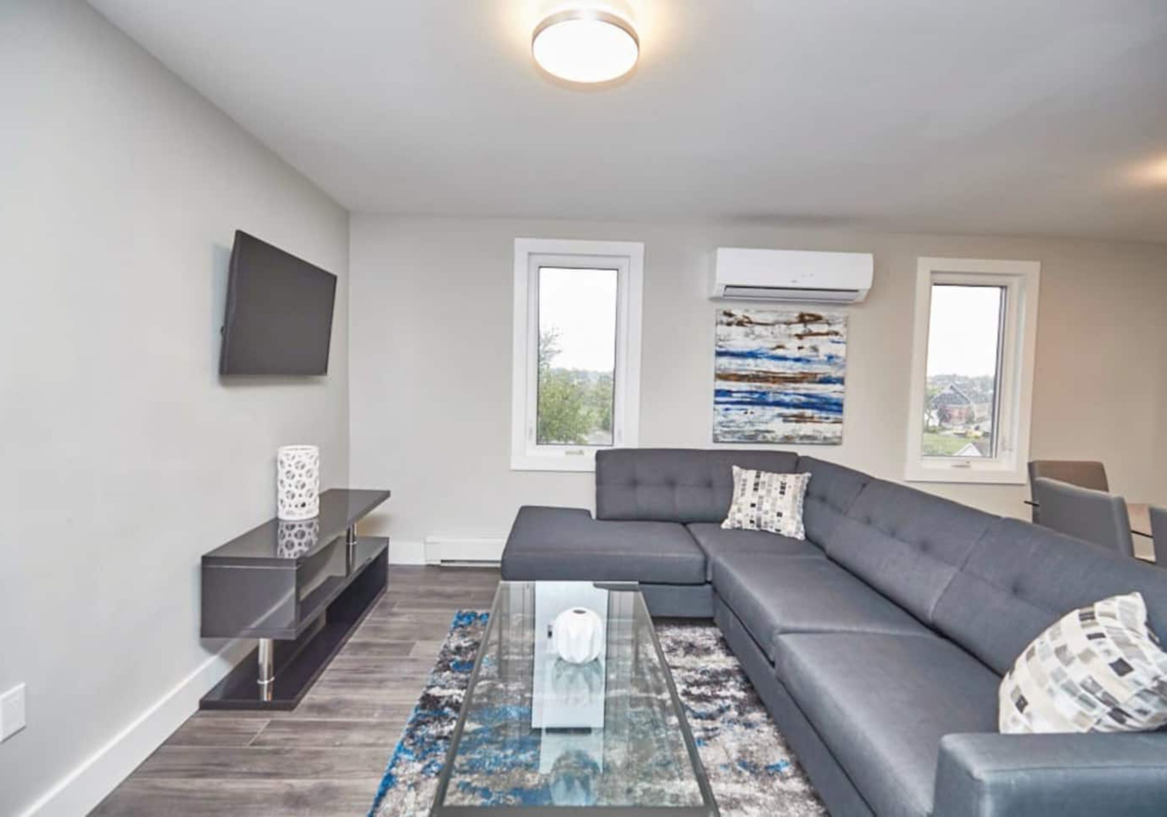 A modern living room with a grey sectional sofa, a glass coffee table, and a flat-screen TV mounted on the wall. There are two windows with air conditioning units above them, and a painting is visible on the wall.