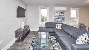 Uma sala de estar moderna com um sofá seccional cinza, uma mesa de centro de vidro e uma TV de tela plana na parede. Há duas janelas com aparelhos de ar condicionado acima, e uma pintura é visível na parede.