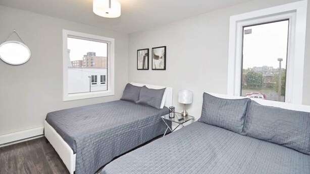 A bedroom with two beds, a bedside table with a lamp, and a window with a view of buildings. - This condo is perfect for families, friend groups and couples. (Niagara Falls)