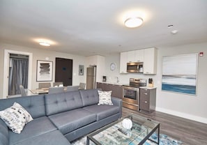 A modern living room with a grey sofa, a coffee table, and a kitchen area with a microwave and oven. - This condo is perfect for families, friend groups and couples. (NIAGARA FALLS)