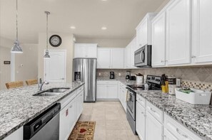 A kitchen with white cabinets, a stainless steel refrigerator, a microwave, and a granite countertop. - Promotion! 8BR Disney Villa | Pool, Spa, Game Room (Kissimmee)