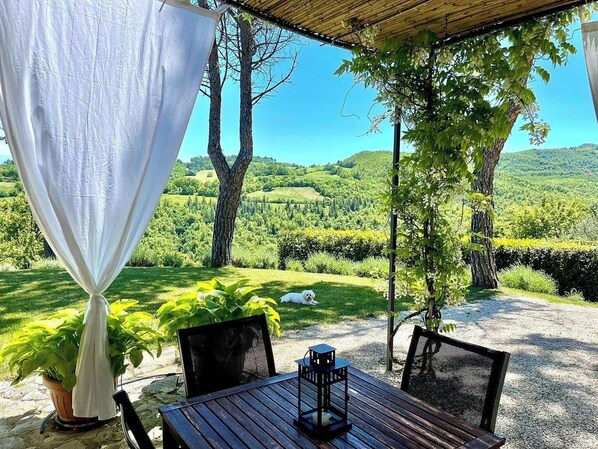 Eine Terrasse im Freien mit Tisch und Stühlen, einem weißen Vorhang und Blick auf eine grüne Landschaft.