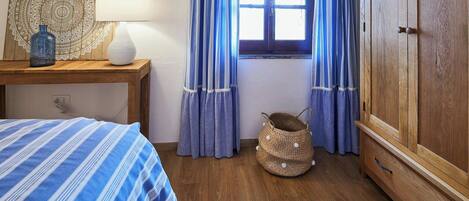 A bedroom with a bed, a bedside table with a lamp, a window with curtains, a wooden wardrobe, and a woven basket.