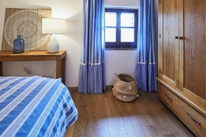 Ein Schlafzimmer mit einem Bett, einem Nachttisch mit Lampe, einem Fenster mit Vorhängen, einem Holzschrank und einem gewebten Korb.