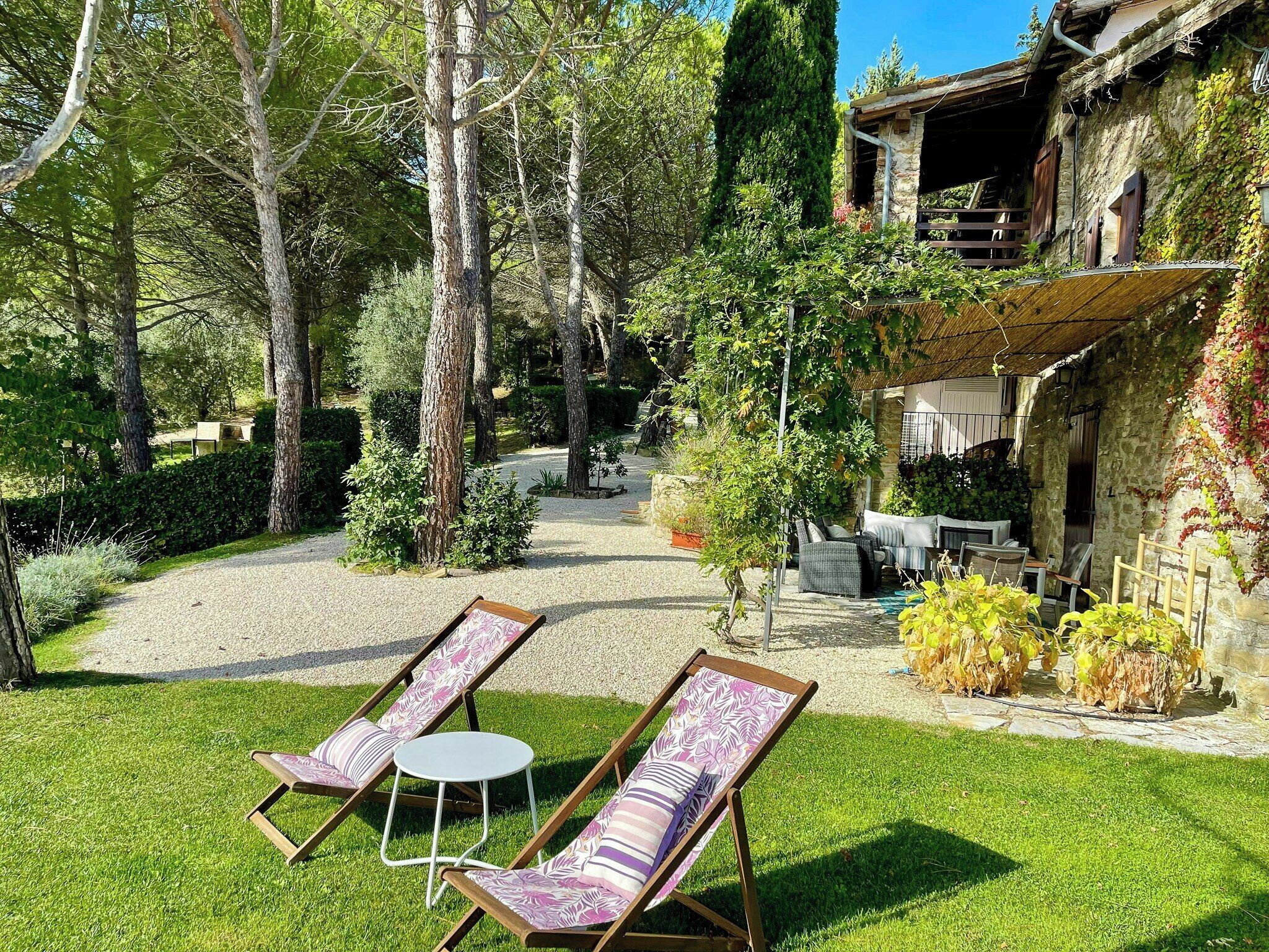 A courtyard with two lounge chairs and a table, surrounded by trees and a stone building.