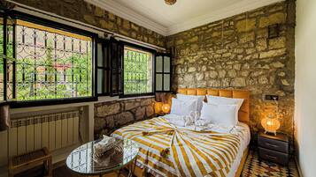 Una habitación con pared de piedra, cama con ropa de cama a rayas, una mesita y una ventana con barrotes.