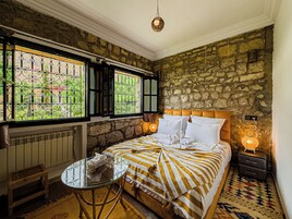 Una habitación con pared de piedra, cama con ropa de cama a rayas, una mesita y una ventana con barrotes.