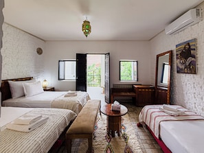 A hotel room with two beds, a bench, a small table, a mirror, and a view of the outside.