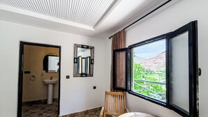 A room with a tiled floor, a wooden chair, a round table, a window with a view of greenery, and a mirror on the wall. - Tonaroz House (Tamatert)