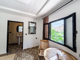 Una habitación con piso de baldosas, una silla de madera, una mesa redonda, un ventanal con vista a follaje y un espejo en la pared.