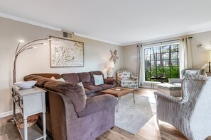 A living room with a brown sofa, a side table, a lamp, a chair, and a large window with a view of greenery. - Seascape Resort Villa | Golden Pond Pool | Minutes to the Beach! (Miramar Beach)