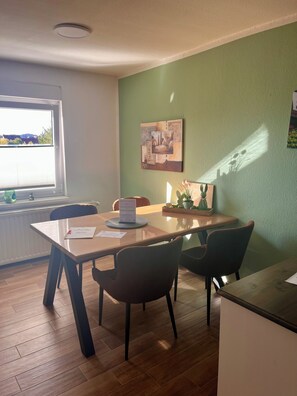 Un coin repas comprenant une table en bois, des chaises, une fenêtre avec des stores, un radiateur et une œuvre d’art accrochée au mur.