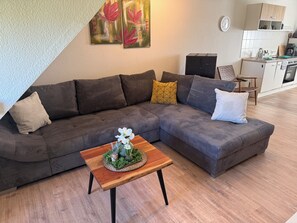 A modern living room with a sectional sofa, a wooden coffee table with a floral arrangement, and a kitchen area in the background. - Westerhäuser Vacation Rentals - Bergzauber Apartment - Hiking in the Harz Mountains (Thale)