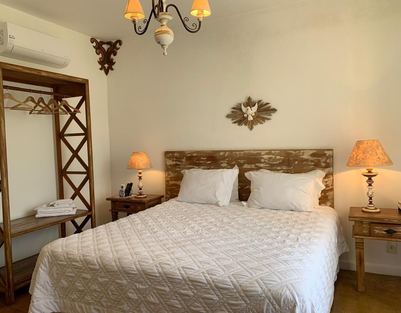 Una cama bien hecha con sábanas blancas, dos mesitas de noche con lámparas, un cabecero de madera, un aire acondicionado montado en la pared y un armario de madera con ropa.