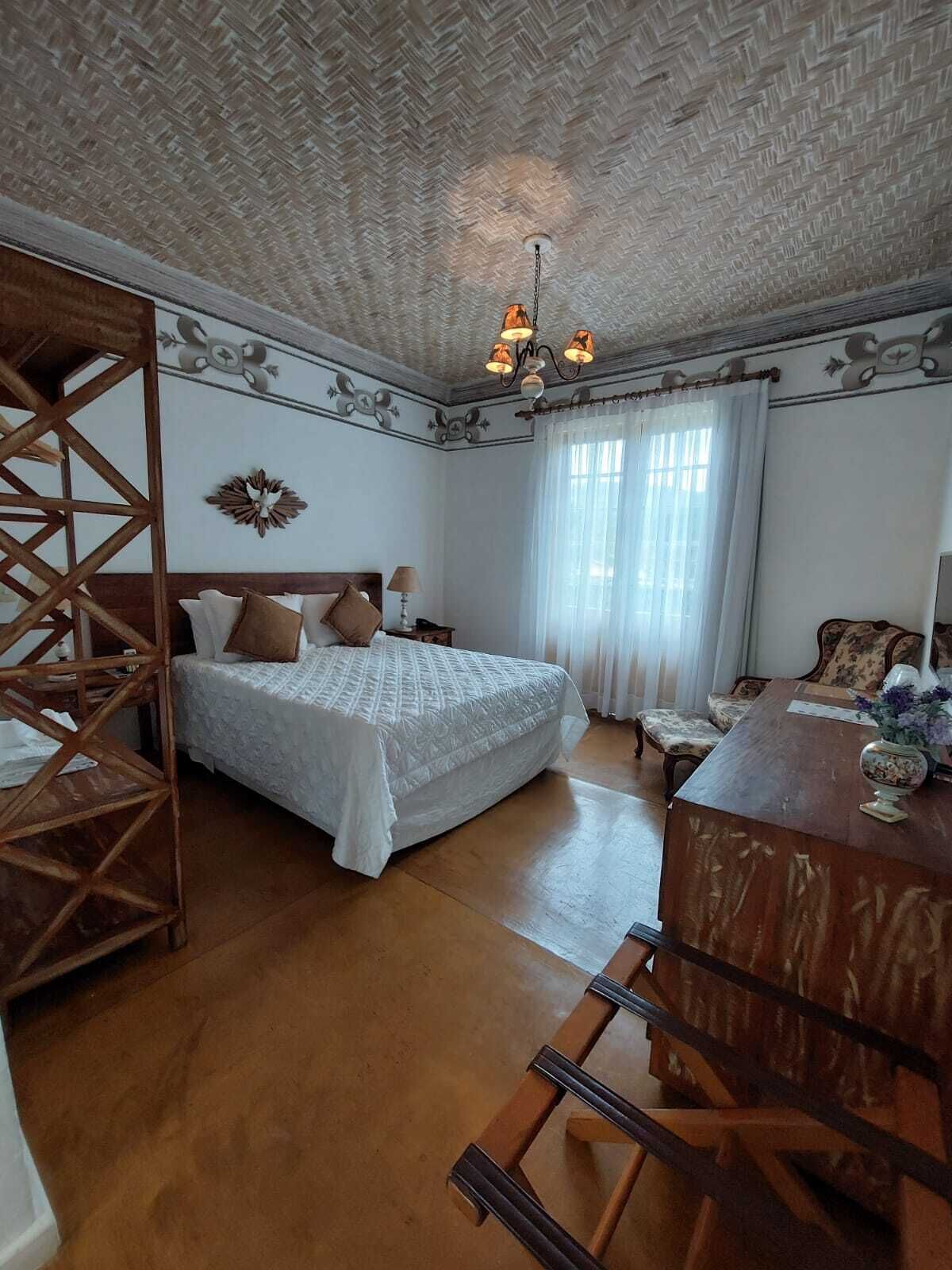 A bedroom with a large bed, a wooden chair, a desk, and a chandelier.