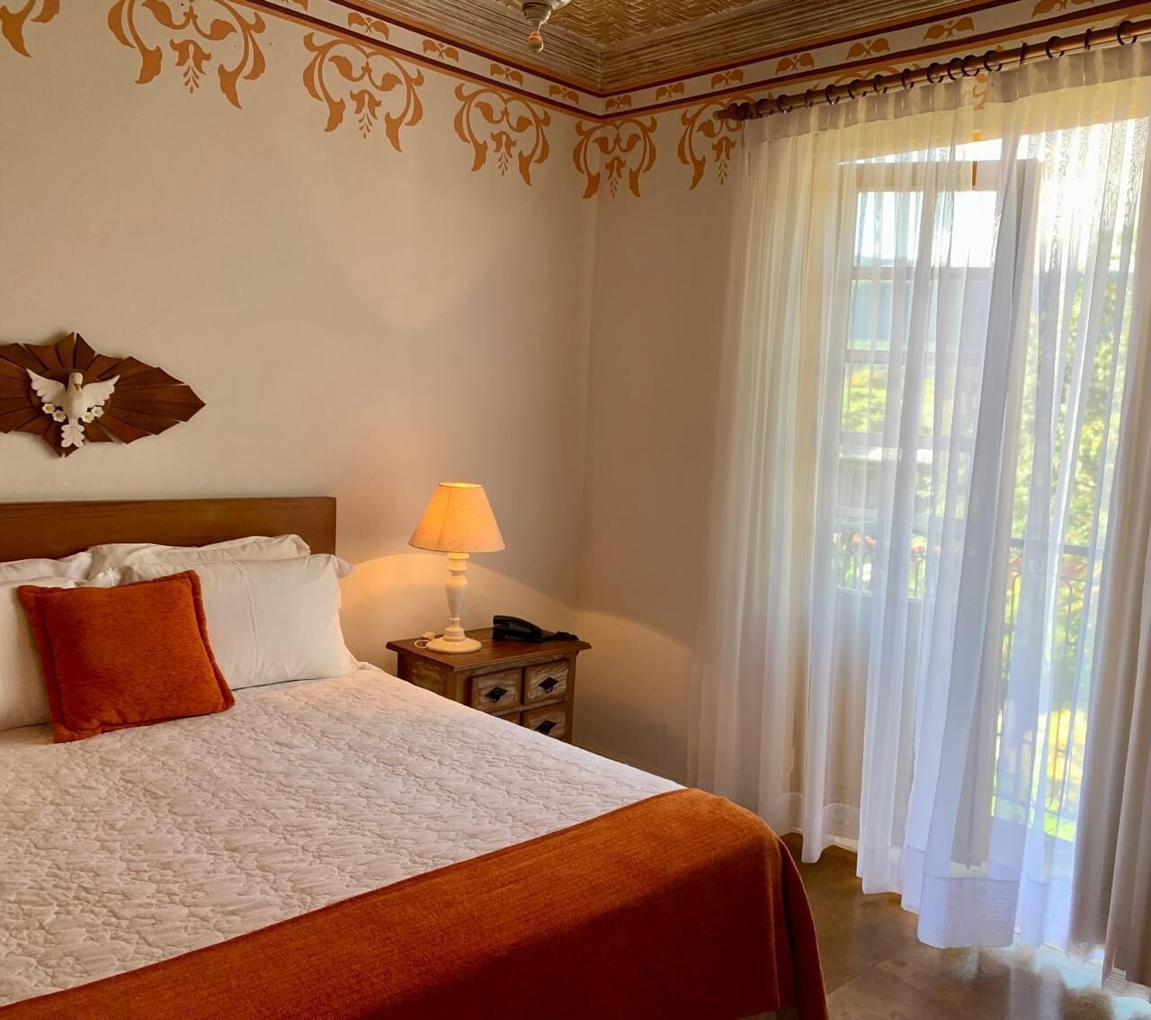 A hotel room with a bed, a nightstand with a lamp, and a view of greenery through sheer curtains.