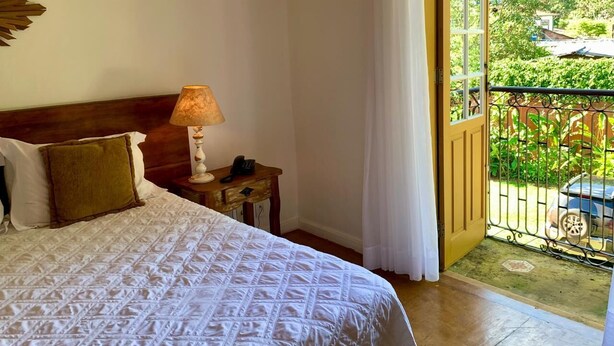 Habitación de hotel con cama, mesita de noche con lámpara, teléfono y vistas a un balcón con plantas y un coche.
